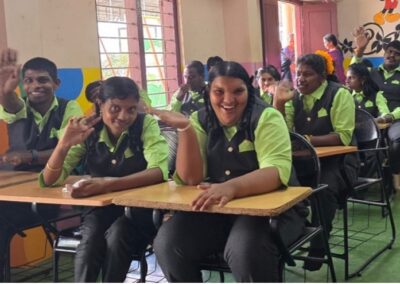 Schülerinnen und Schüler der HOM Special School sitzen lächelnd in einem Klassenraum und winken in die Kamera.