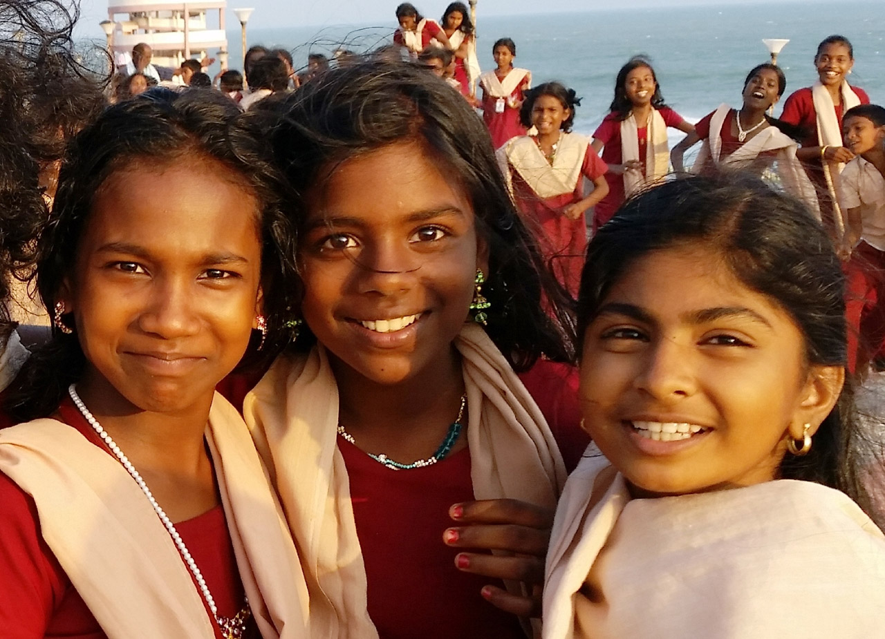 Aus Chancen wächst Zukunft. Drei lächelnde Schülerinnen blicken in die Kamera. Im Hintergrund eine weitere Gruppe strahlender Kinder und blaues Meer.