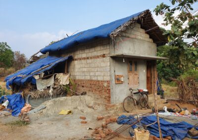 Ein einfaches Wohnhaus mit teilweise provisorisch abgedecktem Dach. Vor dem Haus liegen Baumaterialien, Planen und ein Fahrrad.